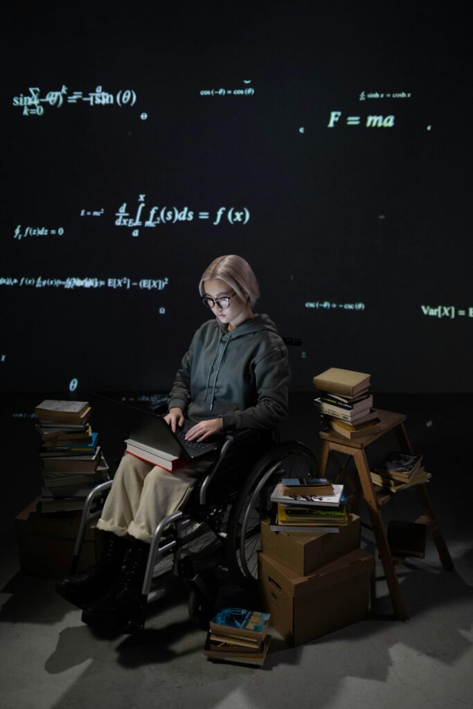 Blonde woman in wheelchair studying math on laptop surrounded by books indoors.